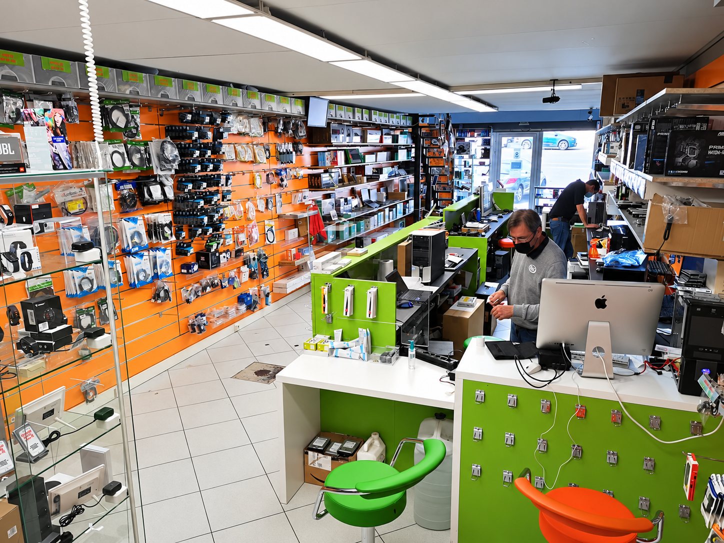 Intérieur d'une boutique d'informatique et d'accessoires avec murs orange couverts de câbles, comptoirs verts et un employé masqué devant un ordinateur.