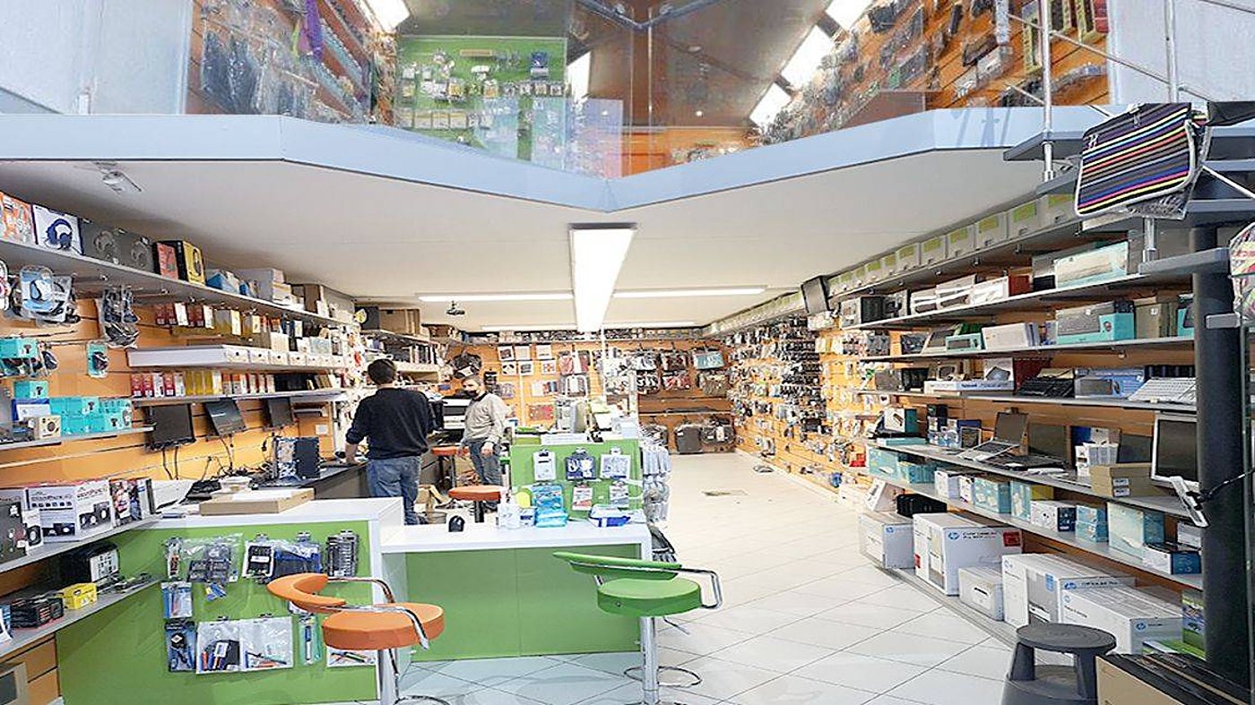 Vue de l'intérieur d'un magasin d'informatique lumineux avec des murs d'accessoires, des boîtes d'ordinateurs, des comptoirs verts et deux personnes au fond.